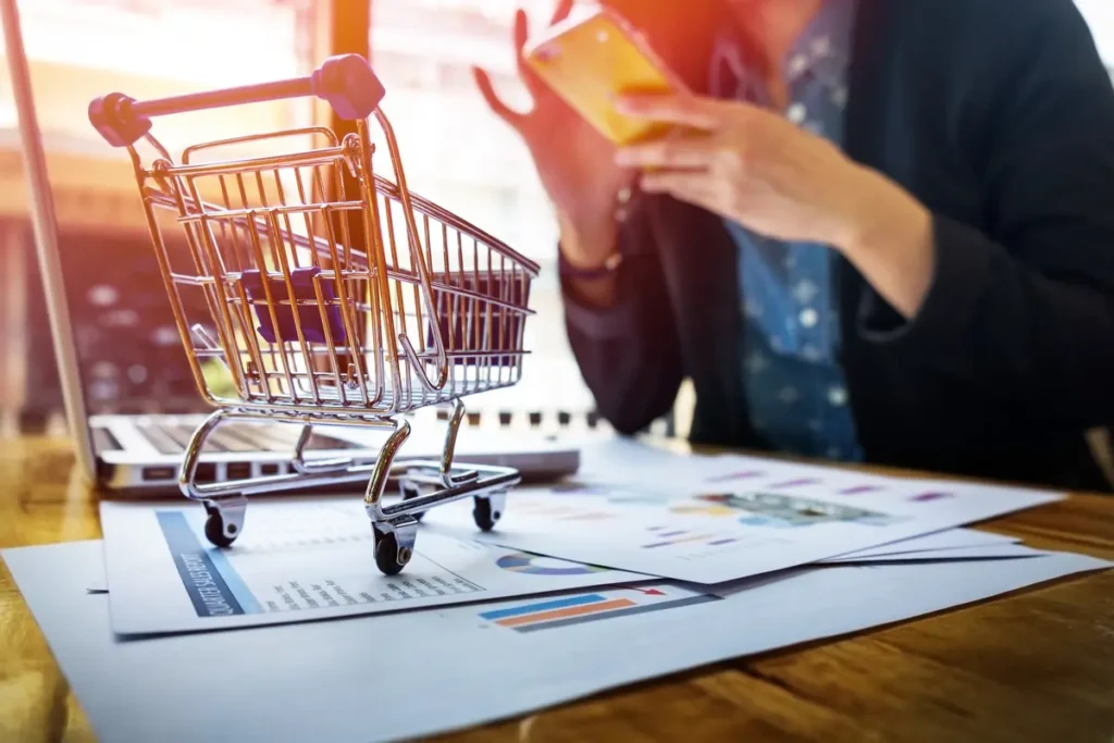 Mini shopping cart placed on business documents with a person using a smartphone in the background, symbolizing mobile e-commerce and digital shopping trends.